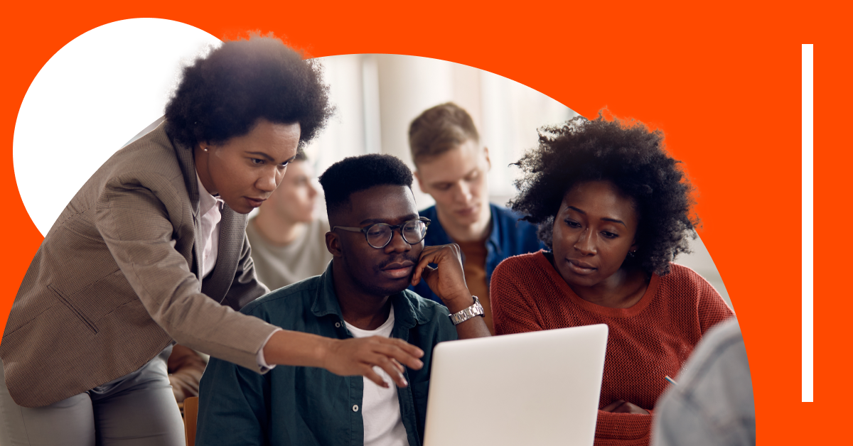 Three people look at a laptop as one person points at the screen; others work in the background. The setting has an orange backdrop with a white graphic element.