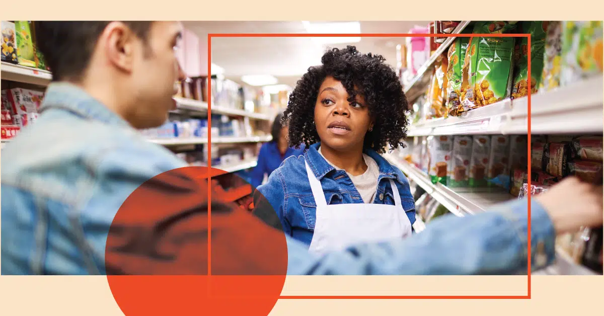 A grocery store employee with curly hair wearing a blue apron speaks to a customer who is reaching for an item on a shelf.