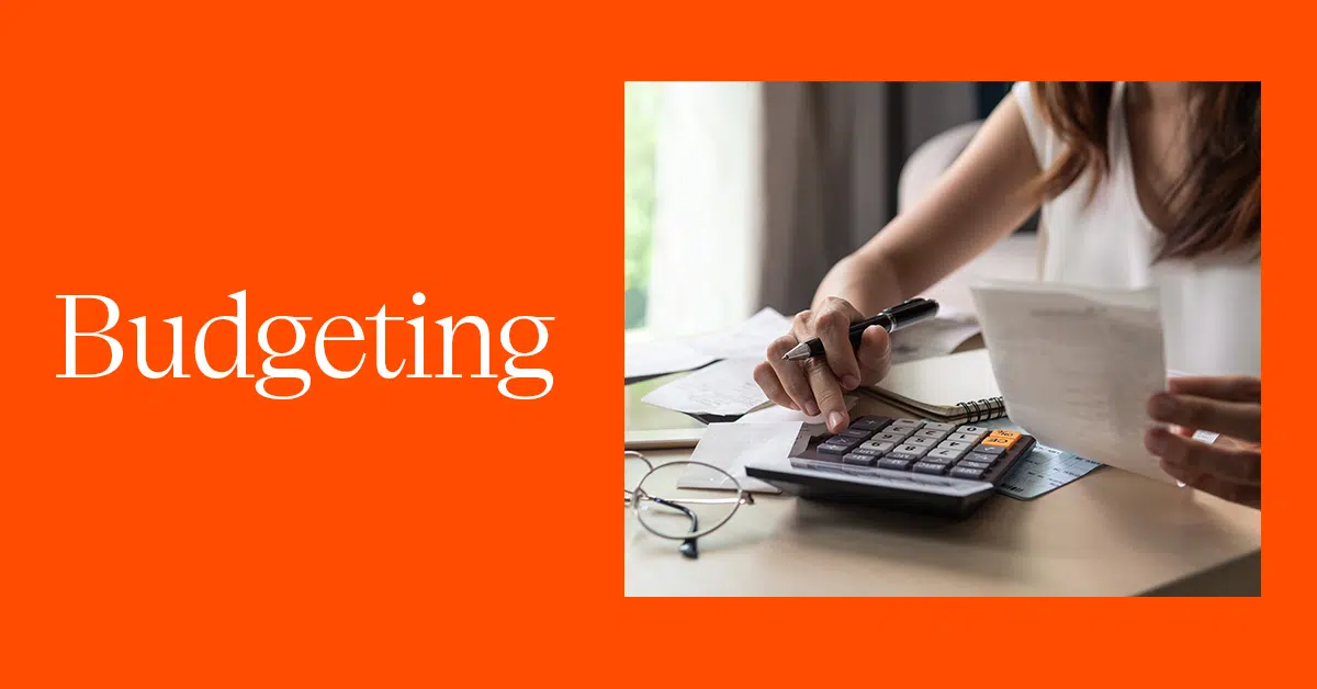 A person sits at a desk with a calculator, pen, and several papers or receipts, seemingly working on finances. The background is a brightly lit room with curtains. To the left, the word "Budgeting" is prominently displayed in white text on an orange background. Glasses rest on the desk.