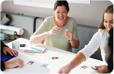 A group of people sitting at a table, smiling and engaging in a card game. There is a notebook, a coffee mug, and scattered cards visible on the table.
