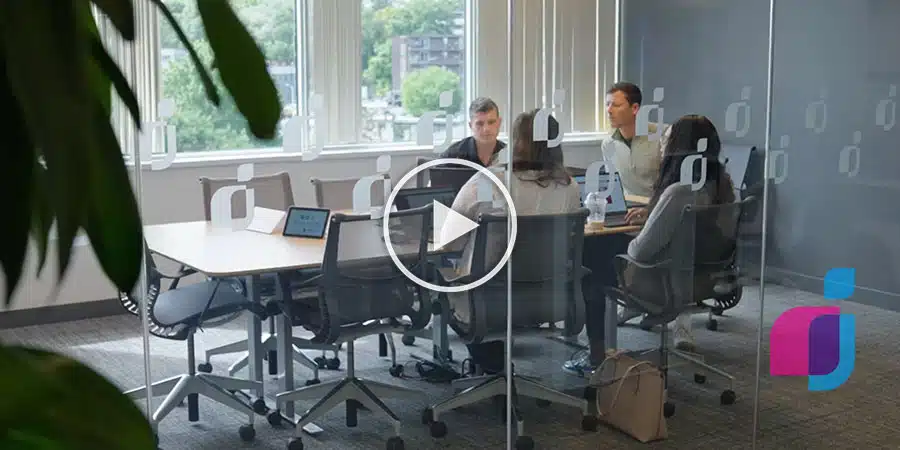 Four people in a meeting room with glass walls, sitting around a conference table. A large screen and laptops are in use. A logo with a blue and purple design is visible in the lower right corner.