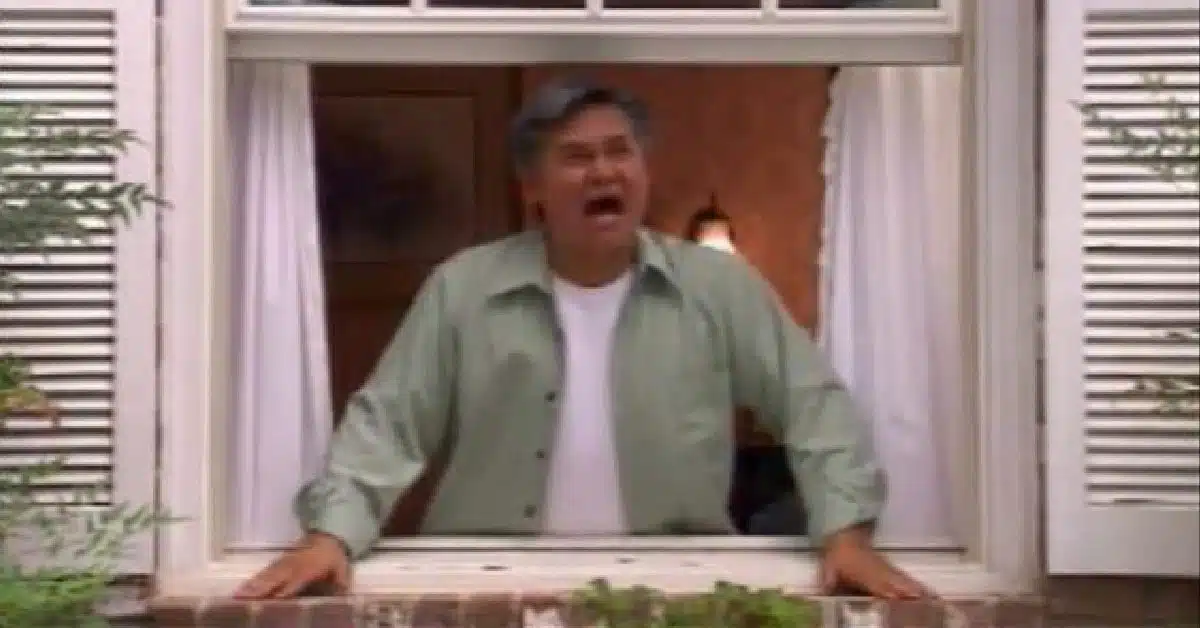 A man with gray hair and wearing a light green shirt is standing at a window, looking outside and shouting.