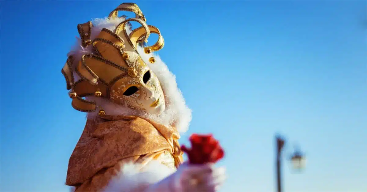 Person dressed in an elaborate gold and white Venetian mask, holding a red rose. The mask features gold embellishments and intricate details, much like the ornate contracts of payday loans. The background shows a clear blue sky with a distant streetlamp.