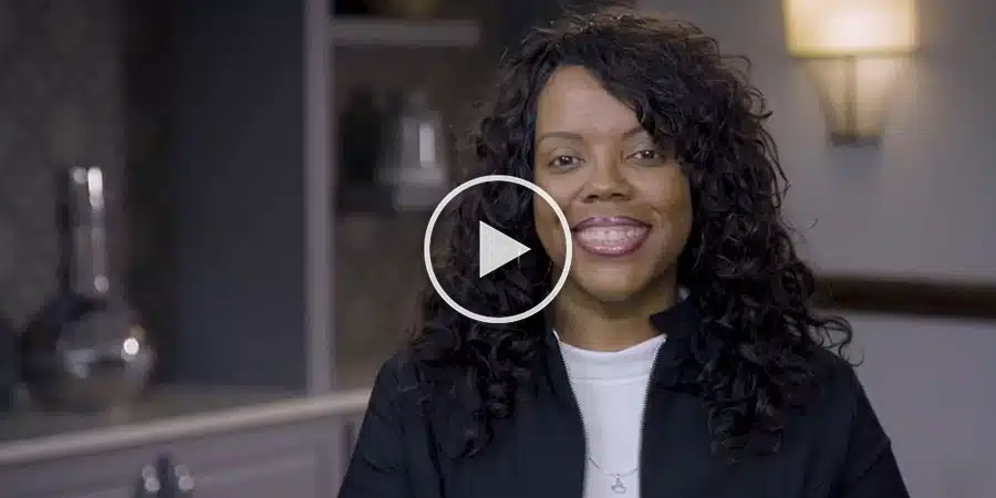 A woman with long curly hair smiles at the camera in a room with a lamp and cabinet in the background. There is a play button overlaid on the image.