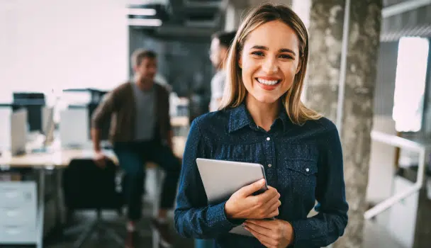 A woman smiles while holding a tablet in an office setting, with two people conversing in the background, depicting a positive work atmosphere that helps reduce employee attrition.