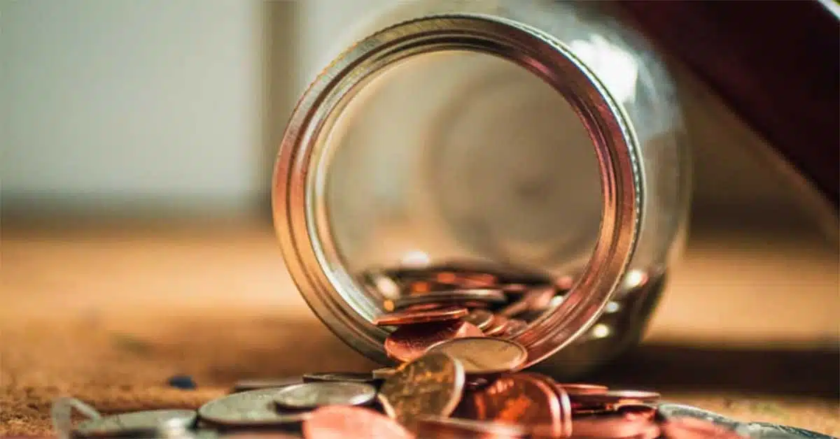 A glass jar lying on its side with various coins spilling out onto a wooden surface, reminiscent of borrowing from your 401K. The coins, primarily copper and silver-colored, scatter in different directions. The background is softly blurred, putting the focus on the jar and the coins.